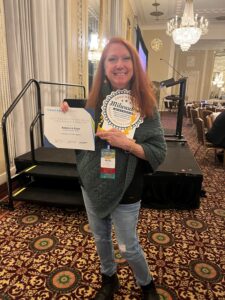 Becky Ryan of the University of Wisconsin-Madison standing and displaying her award at the NACADA Region 5 and 6 conference in March 2024 in Milwaukee 