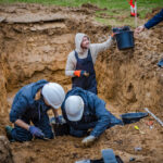Photo of work at the site of a mass grave in Normandy, France, in December 2023. UW Oshkosh associate professor Jordan Karsten hands off a bucket containing bones to be submitted for DNA analysis. Photo courtesy Anibas Photography