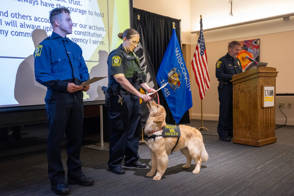 Photo of Ezmae accepting a treat from her handler, UW-Milwaukee Police Officer Mindy Wucherer, during a swearing-in ceremony as Interim Police Chief Brian Switala says a few words. At left is new officer Grayson Smith. (UWM Photo/Troye Fox)